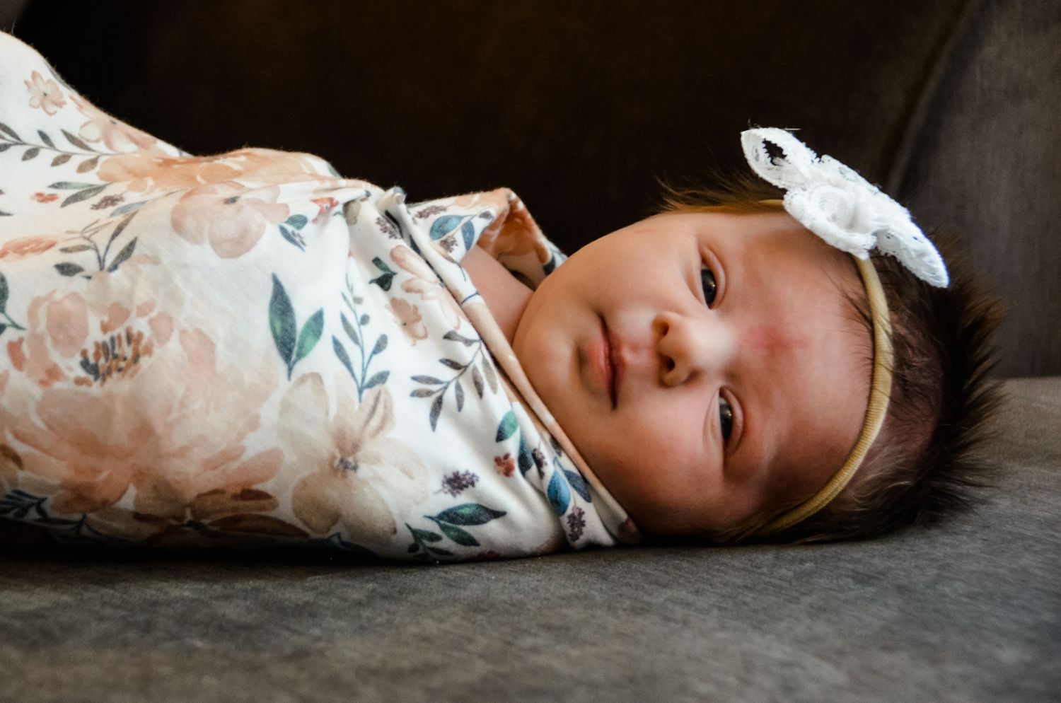 Newborn girl swaddled in floral blanket, wearing a headband with a white bow, with calm expression.
