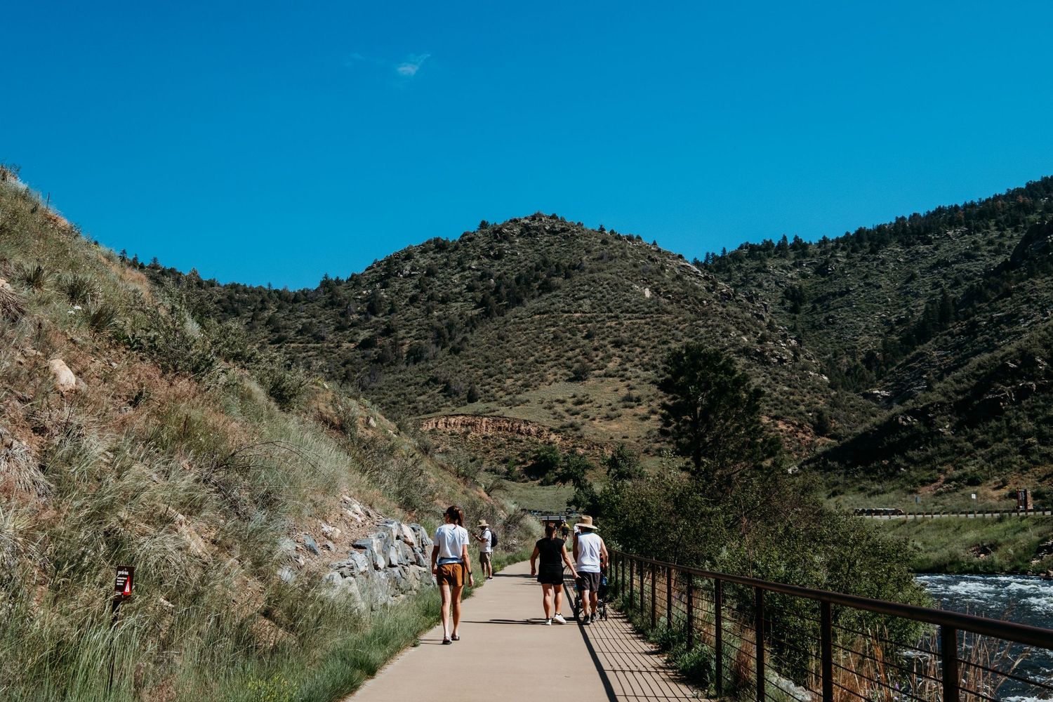 Group hike at Clear Creek Trail, paved path, railing to the right of hikers, river down below, with open blue skies.