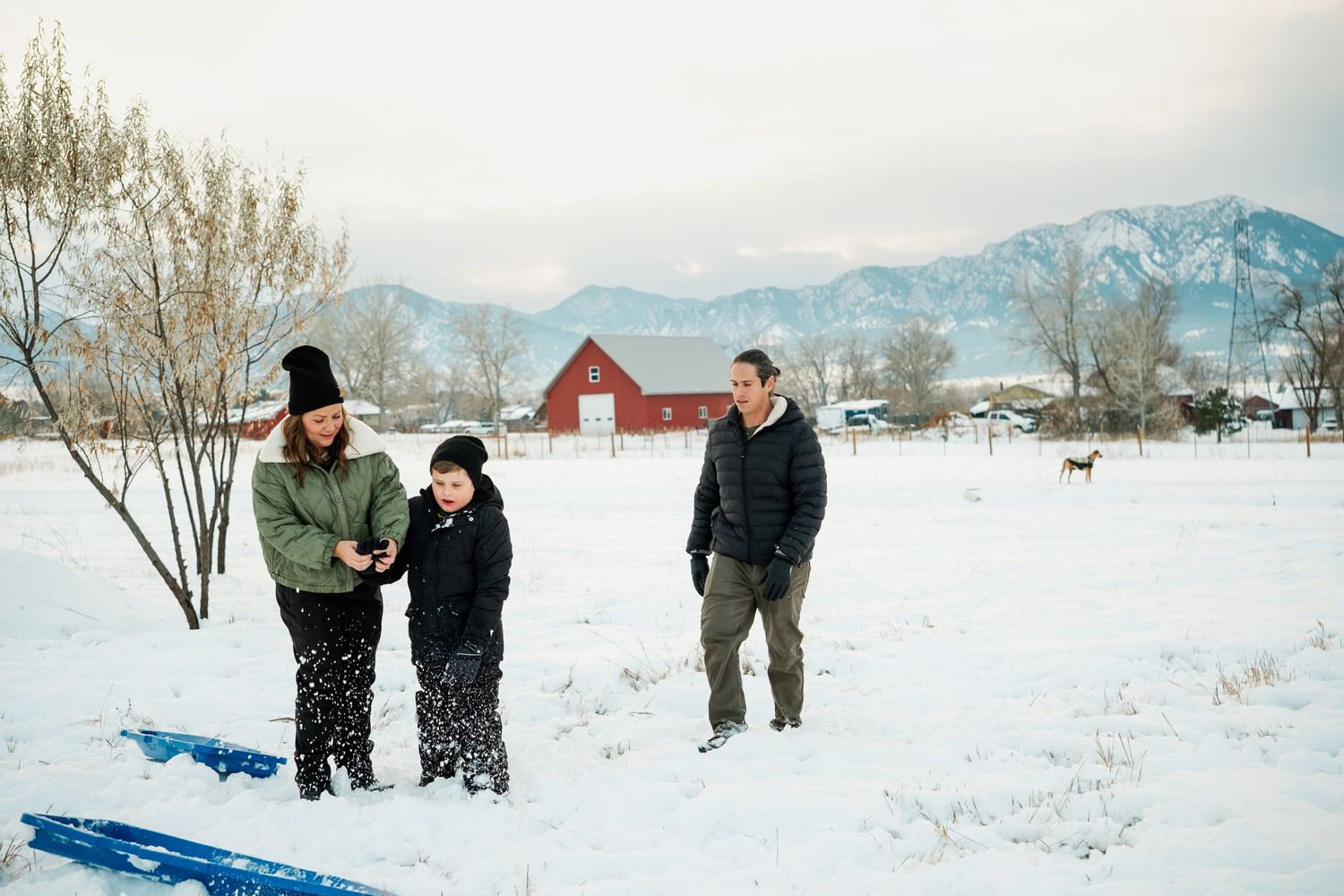 Family of mother, father and young son standing in the snow, a red barn and Boulder flat irons in the background.