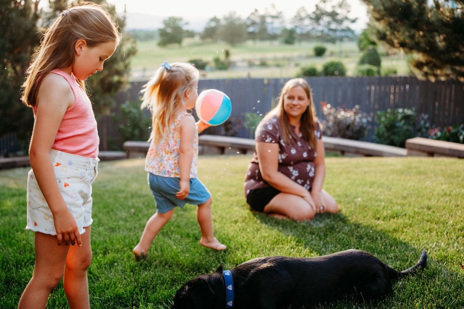 Children walking in backyard, woman sitting in grass in background.
