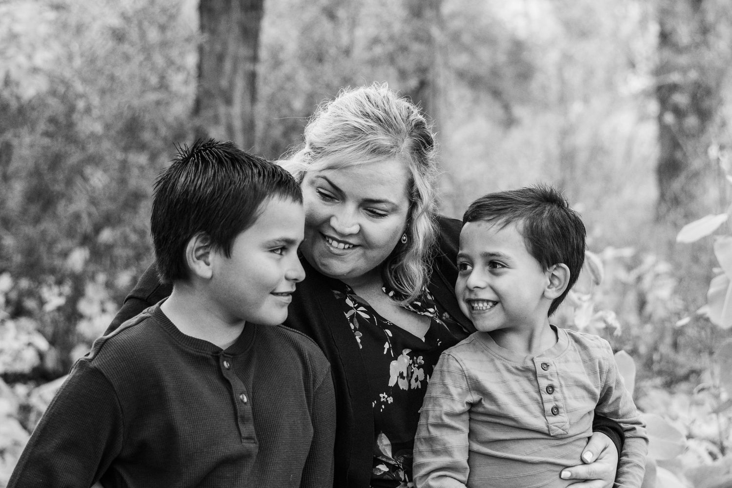 Mother sitting between two sons, all smiling at one another, trees in background.