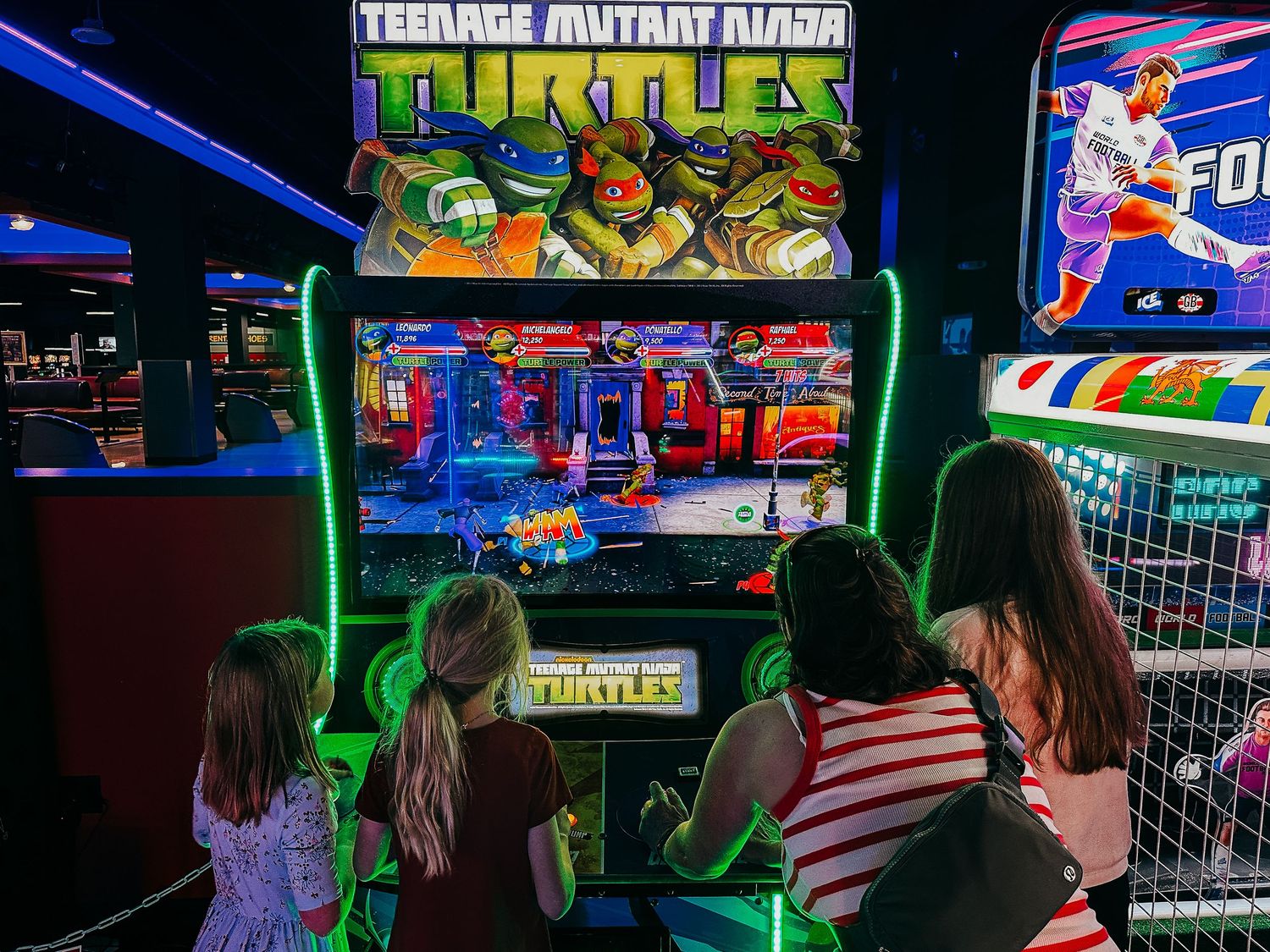 One adult woman with three girls playing Ninja Turtles Arcade game in low light, with vibrant neon colors of the game.