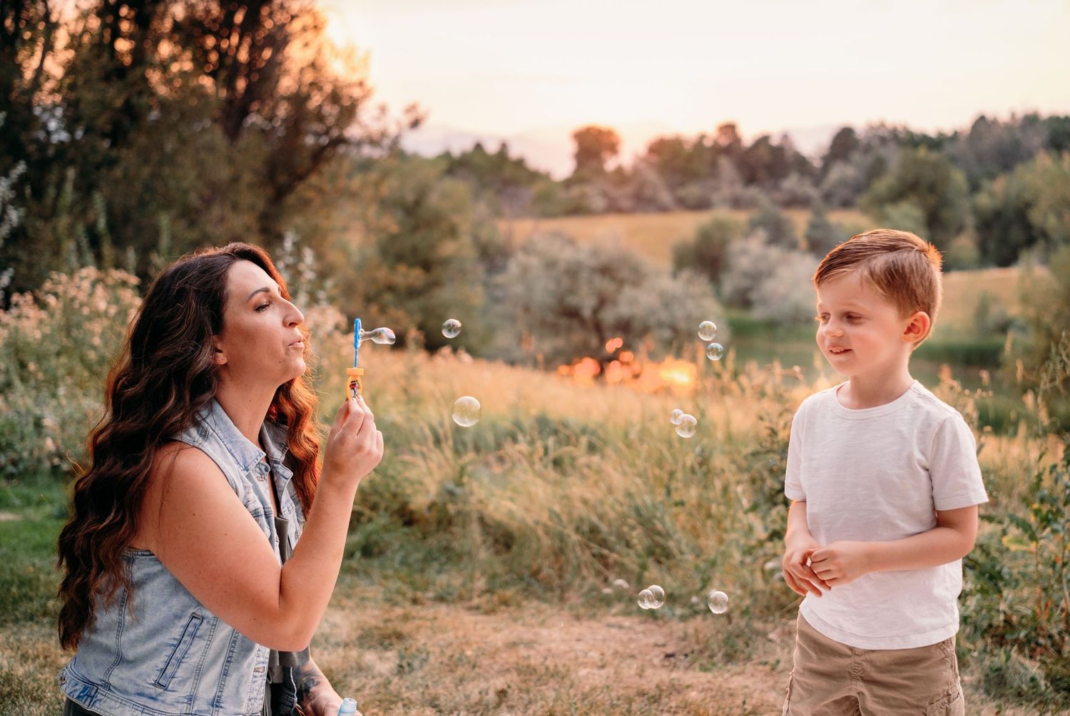 Mother blowing bubbles at son, lake and sunset in background.