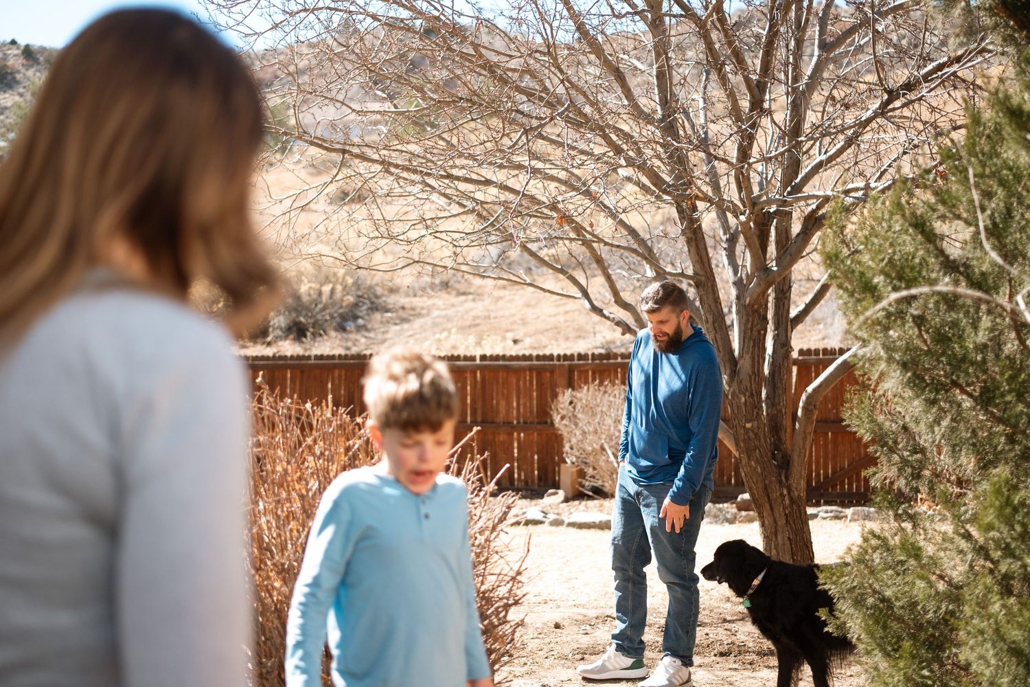 Mother, son and husband in the backyard with their big black dog and a large tree in the bckground.