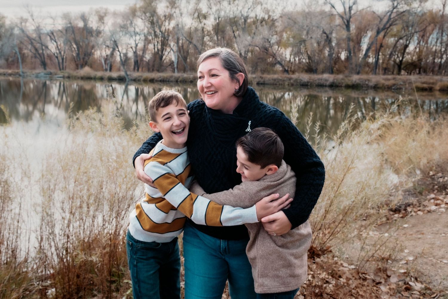 Mother standing as two songs hug her on each side, all three smiling, Golden Ponds Park in the background.