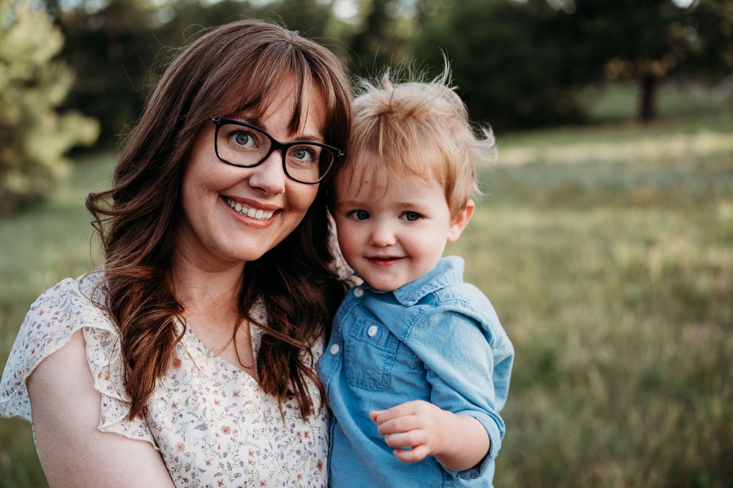 Mother holding toddler son closely, blurred forest background.