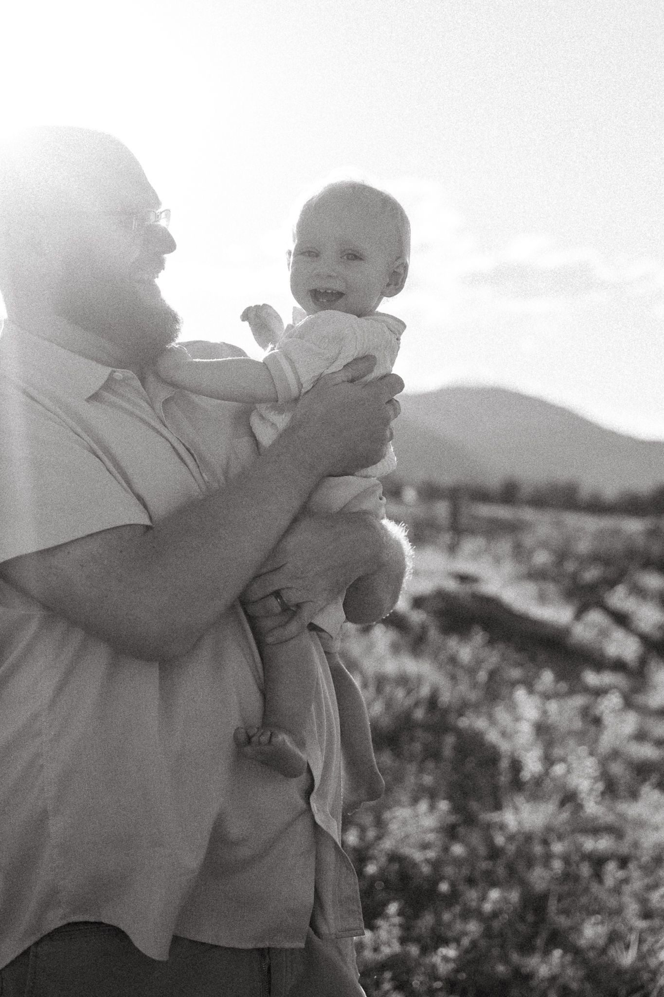 Black and white portrait of father holding one year old son, sun flare backlighting father.