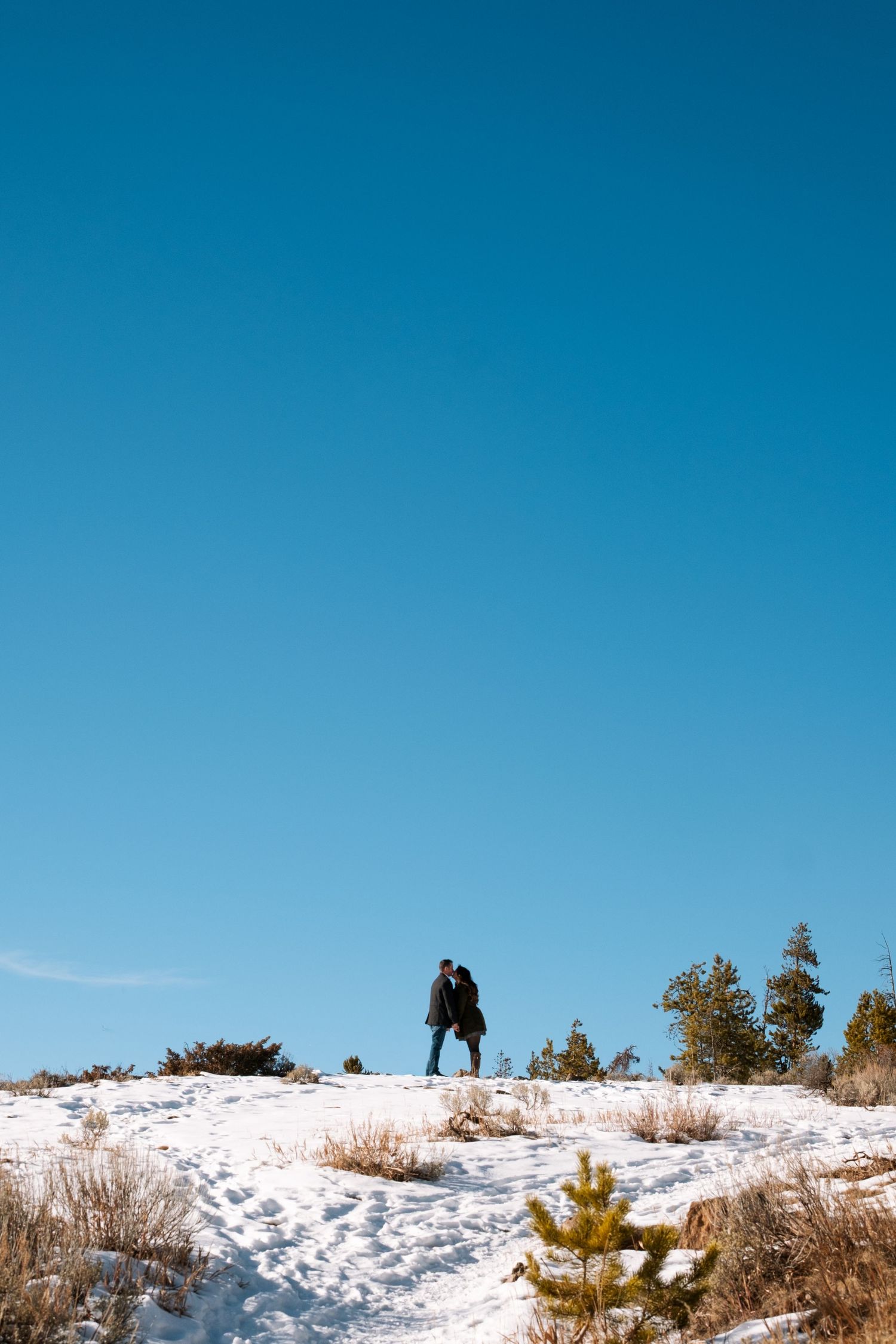 Couple standing on snowy bank, silhouetted by bright blue skies near Breckenridge, Colorado during engagement photo session.