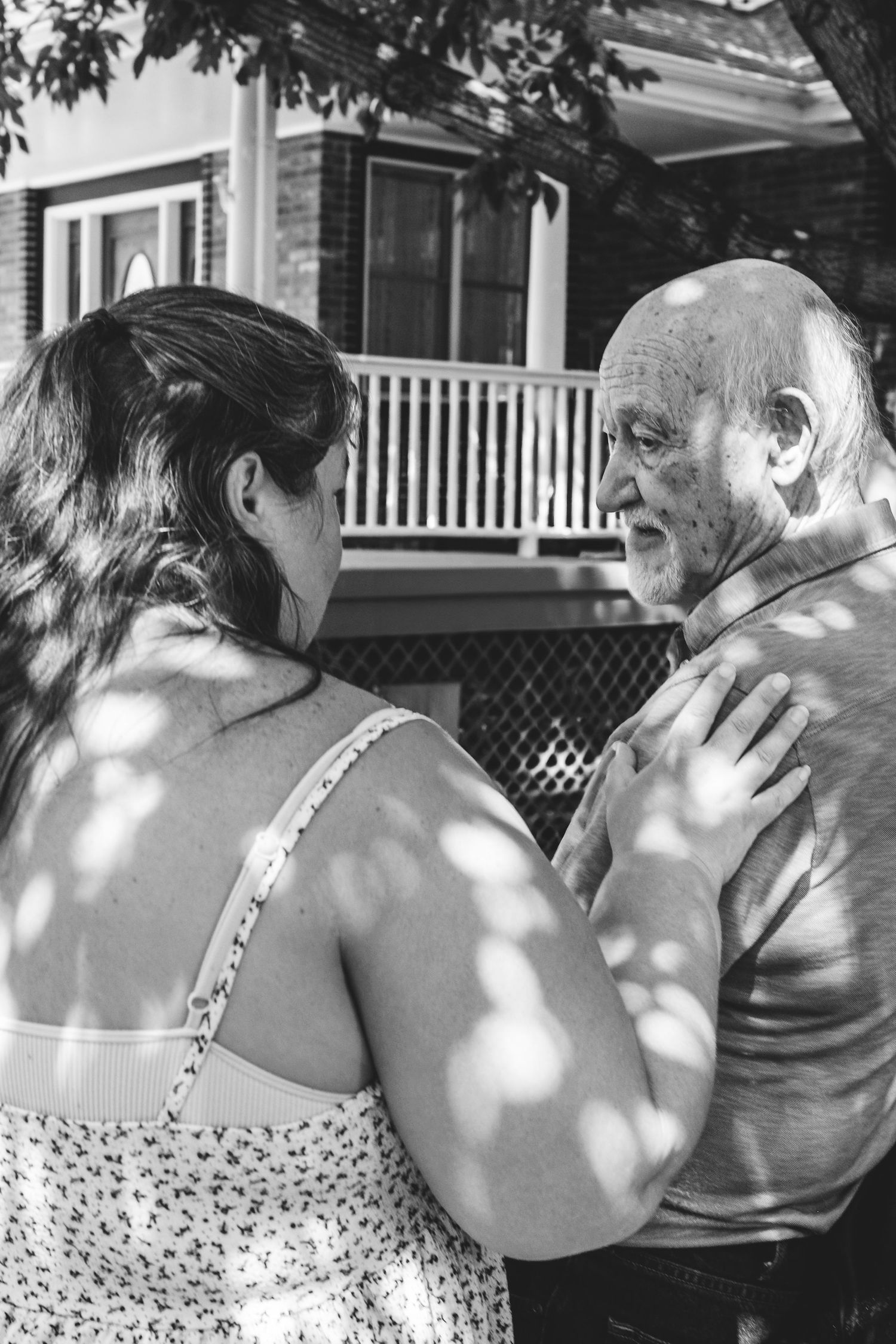 Black and white portrait of adult granddaughter resting hand on grandfather's shoulder, dappled light and shadows.