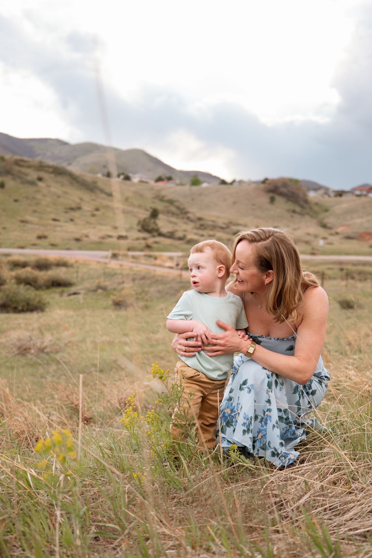 Mother kneeling down low to son in field, tall grass in foreground, dramatic clouds in sky.