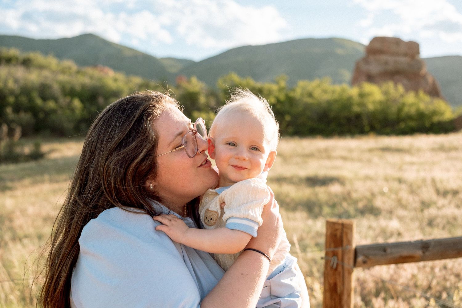 Mother kissing son on the cheek, green shrubs and rock formation in background, at South Valley Park in Littleton, Colorado.