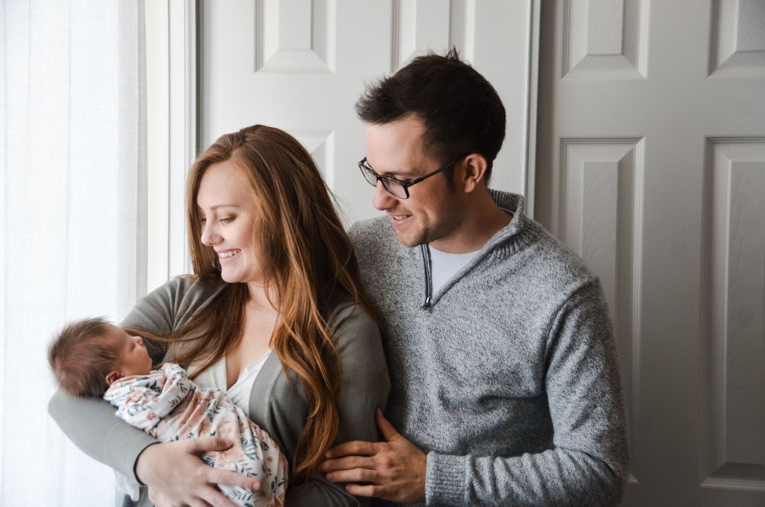 Mother holding newborn daughter, father standing behind mother, both have dreamy lighting from nearby window.