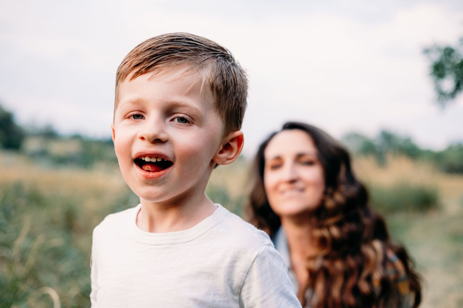 Close up portrait of toddler son, mother gazing toward him in background.
