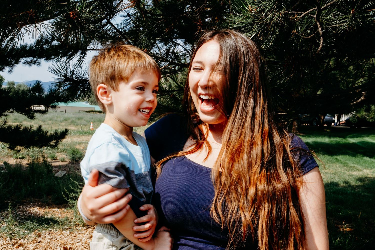 Teen girl laughing while holding toddler cousin, evergreen trees in background.