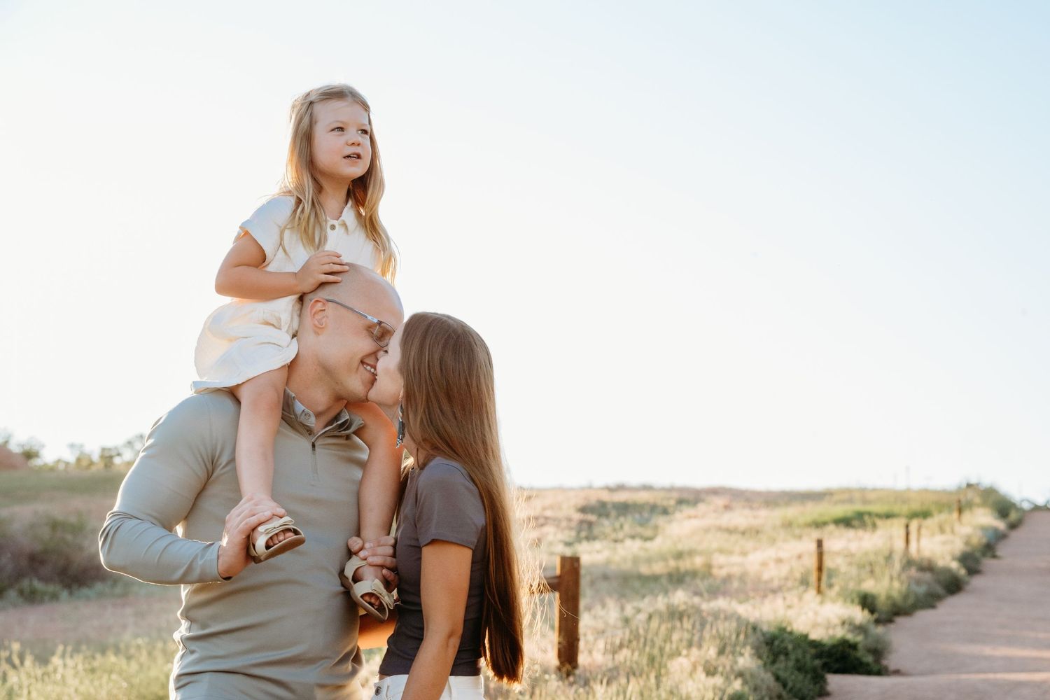 South Valley Park Mini Session with parents and young girl, parents about to kiss, girl perched on father's shoulders, backlit sun.
