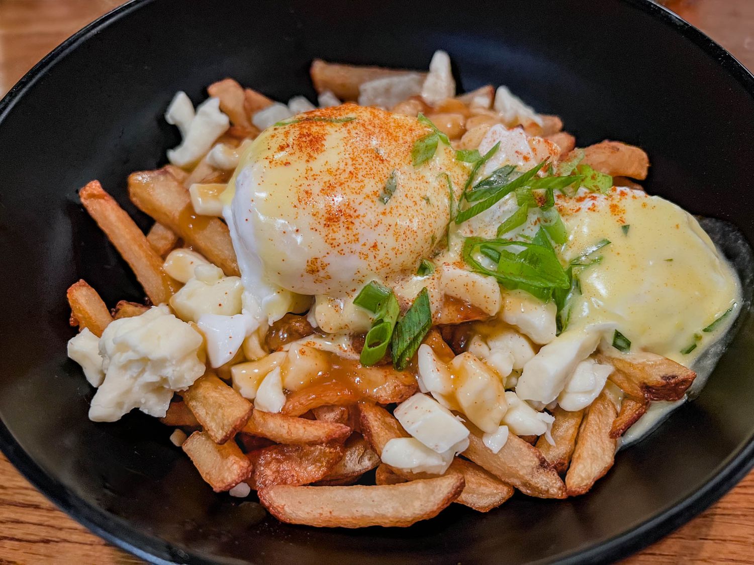 Golden poached eggs with Hollandaise sauce served over crispy french fries and cheese curds in a black bowl.