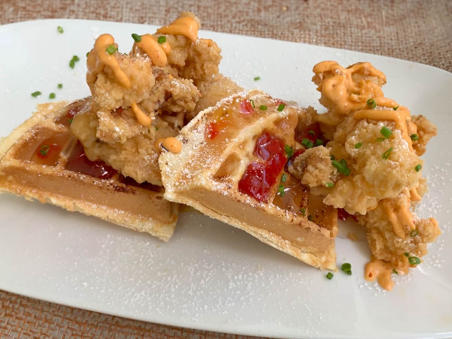 Crispy fried chicken sits atop a golden waffle with red sauce, garnished with fresh herbs on a white plate.