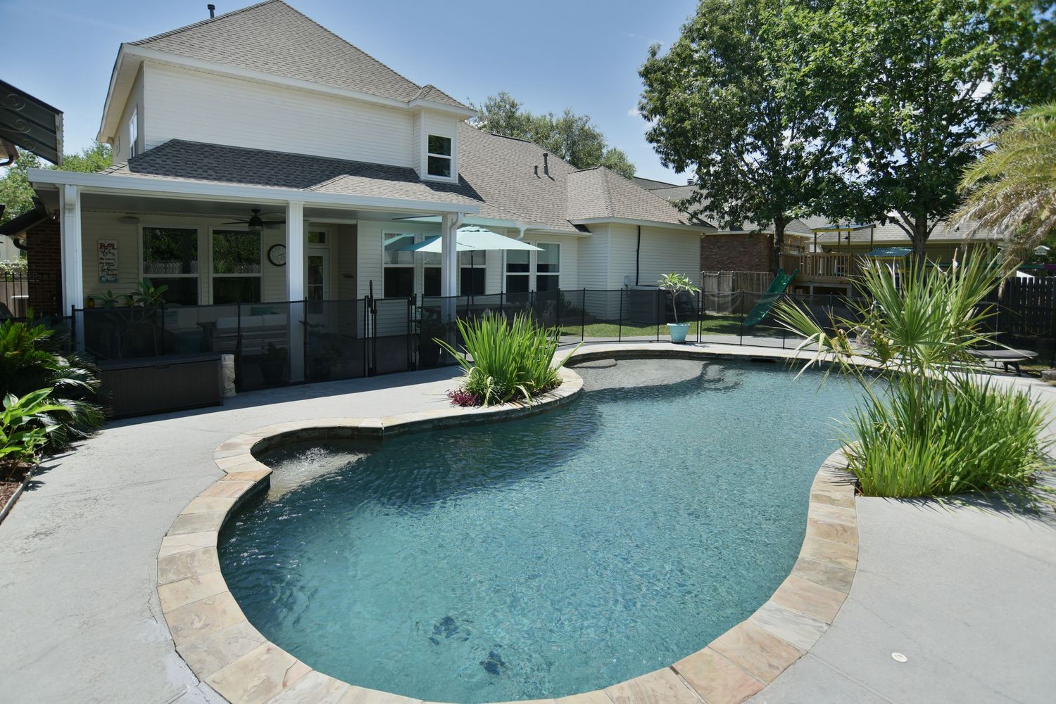 Curved freeform swimming pool with stone coping surrounded by landscaping and covered patio in backyard.