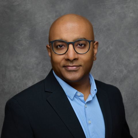 Professional corporate portrait of a business professional in glasses wearing a suit jacket and blue shirt.