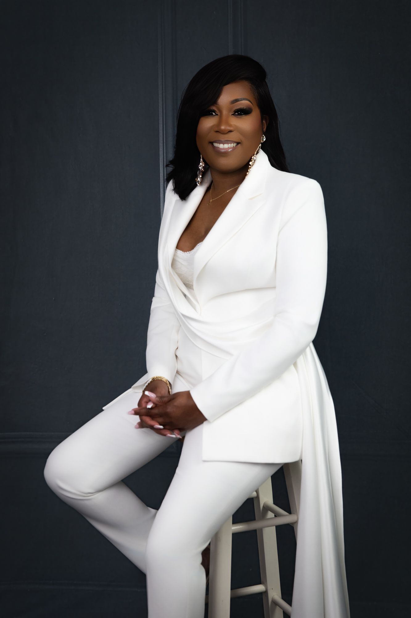 Professional studio portrait of someone in an elegant white pantsuit posing gracefully on a stool against a dark background.