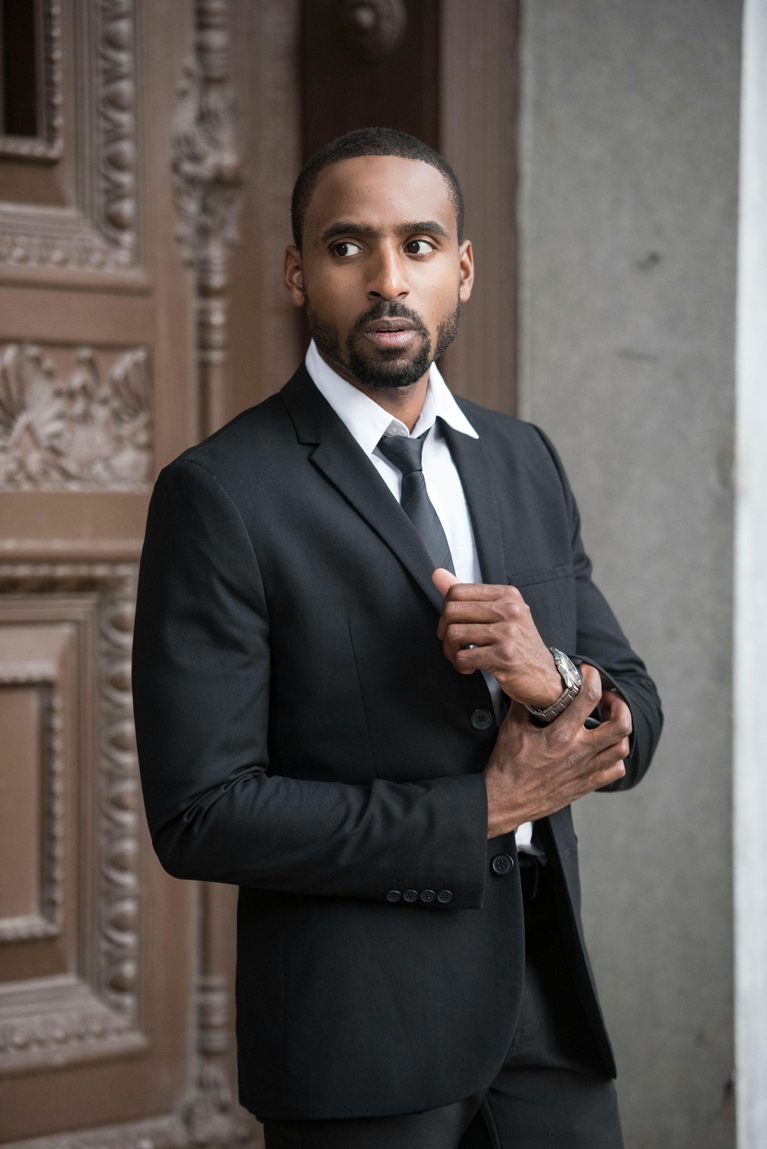 Professional portrait of a well-dressed person in a black suit and tie adjusting their cuff against an ornate doorway.