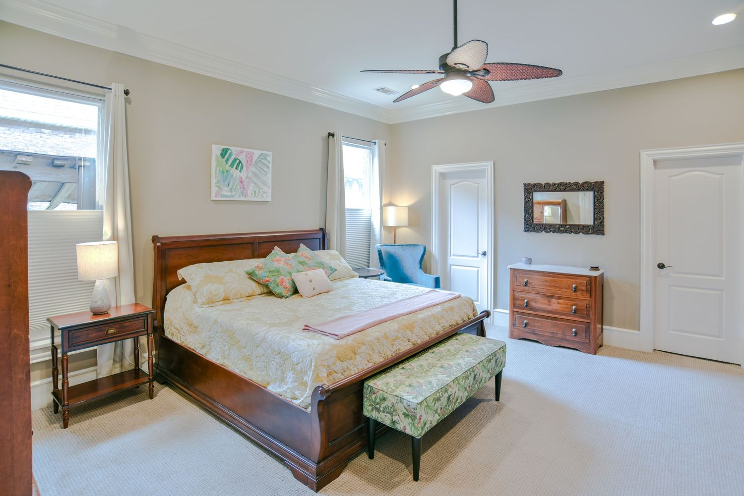 Spacious master bedroom with wooden furniture, beige walls, upholstered bench, and ceiling fan in natural light.