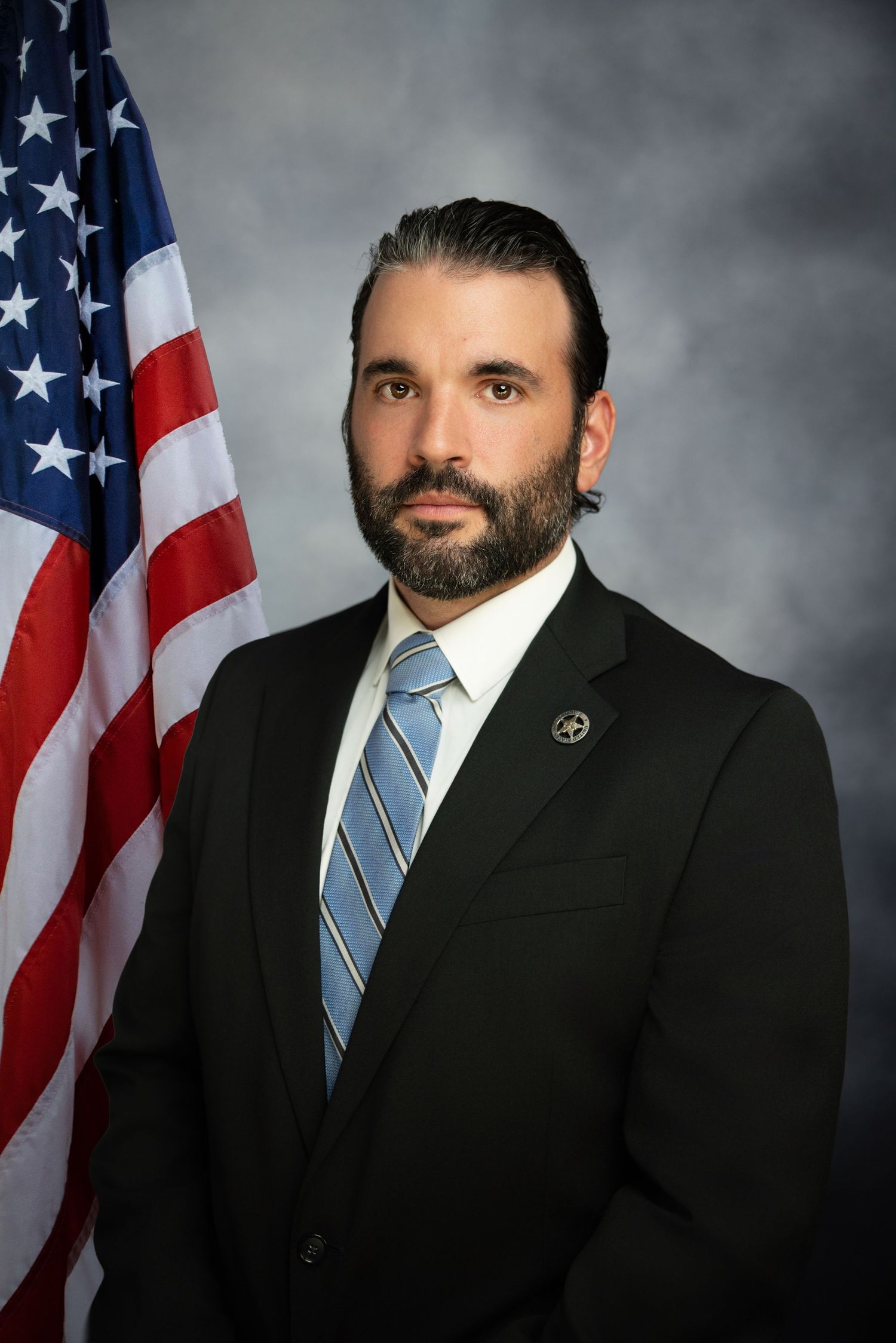Professional headshot portrait of someone in a dark suit and blue striped tie against American flag backdrop.