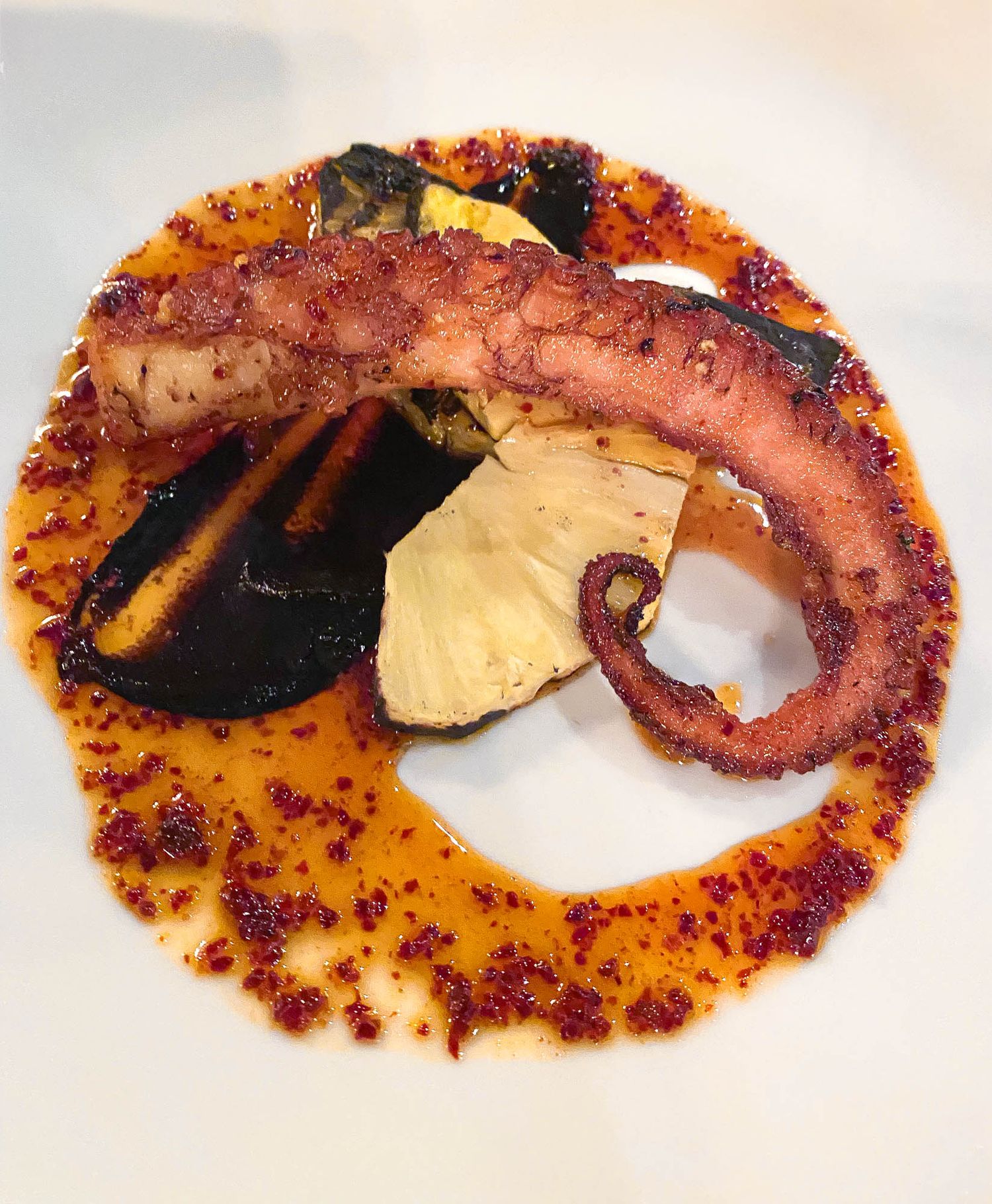 Grilled octopus tentacle served with black mussels and potatoes in a vibrant orange paprika sauce on a white plate.