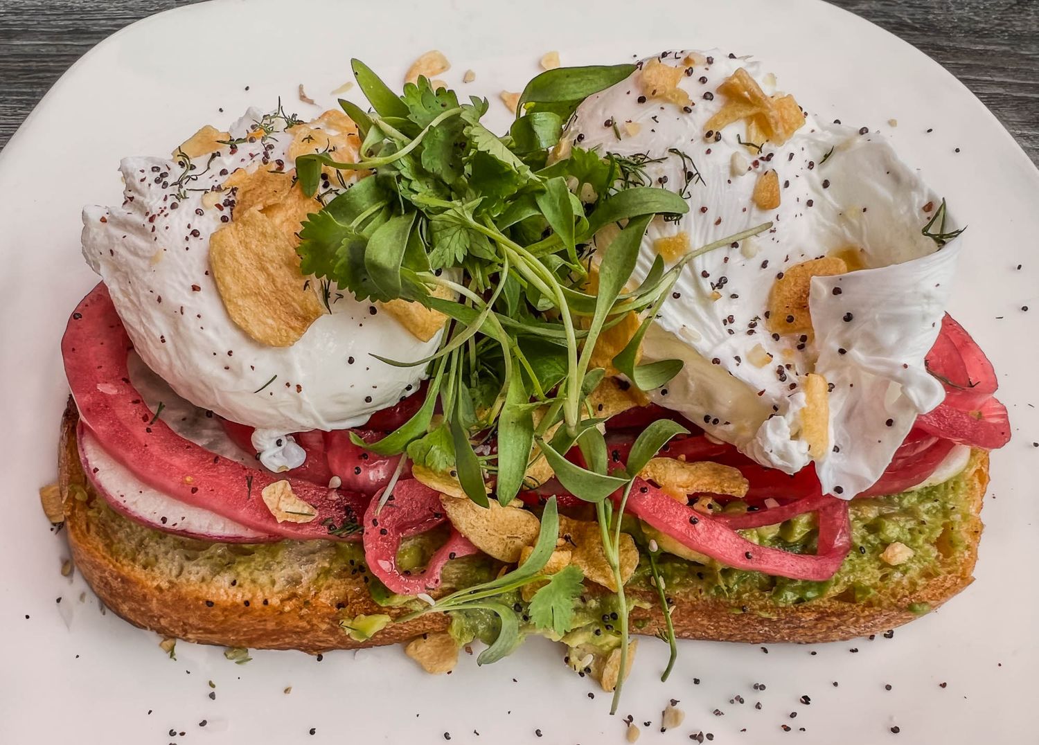 Open-faced sandwich with sliced meat, poached eggs, arugula, and creamy sauce on artisanal bread on white plate.