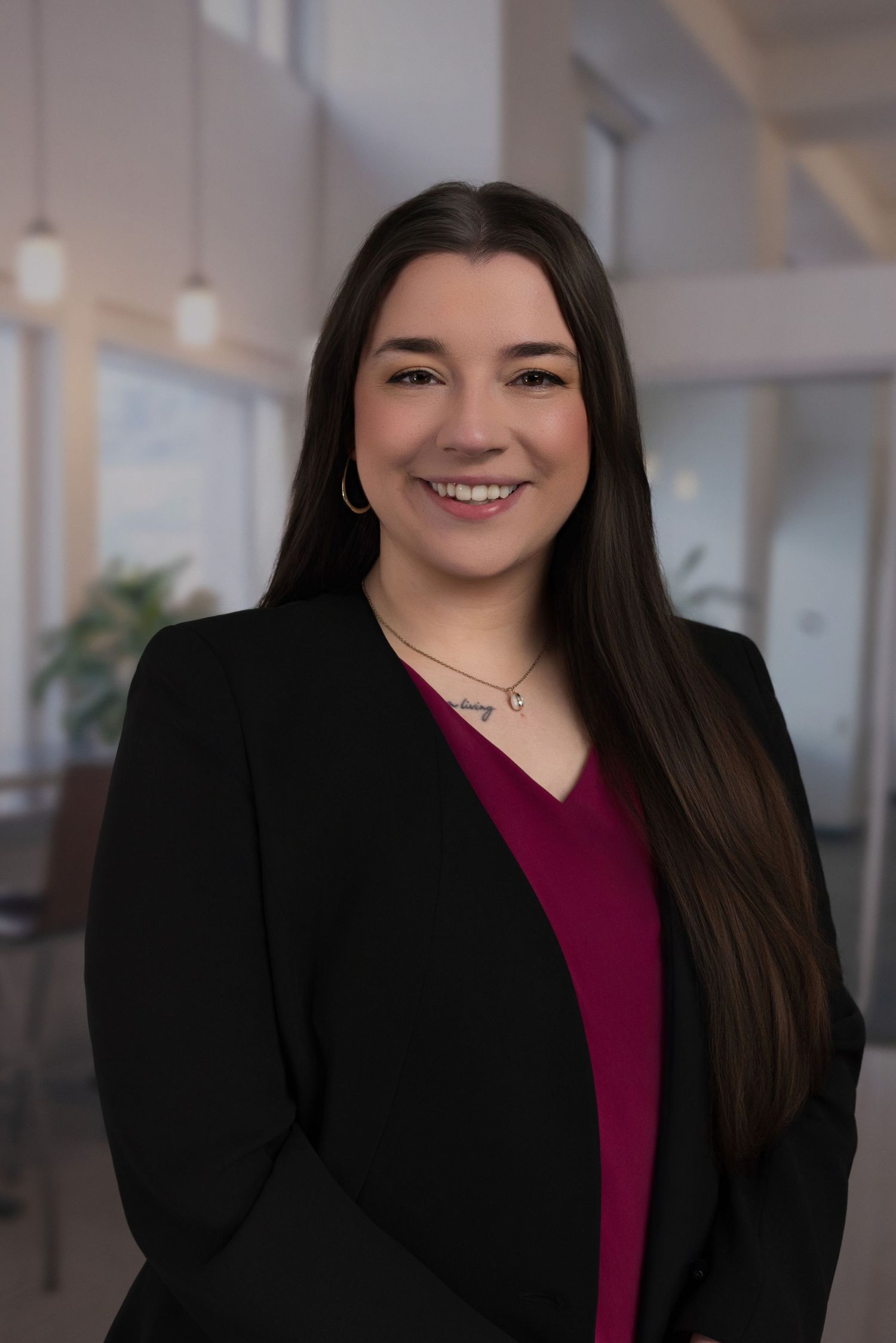 Professional woman with long dark hair smiling in black blazer and magenta top in modern office setting.