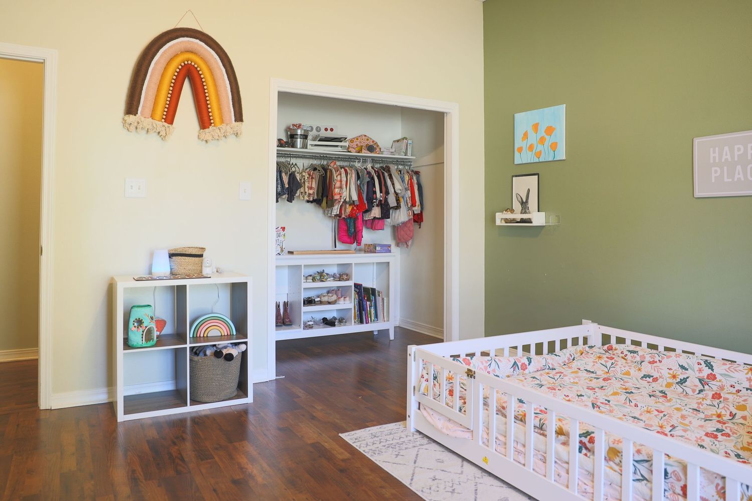 A cozy nursery room with rainbow wall art, built-in closet storage, and a white toddler bed on hardwood floors.