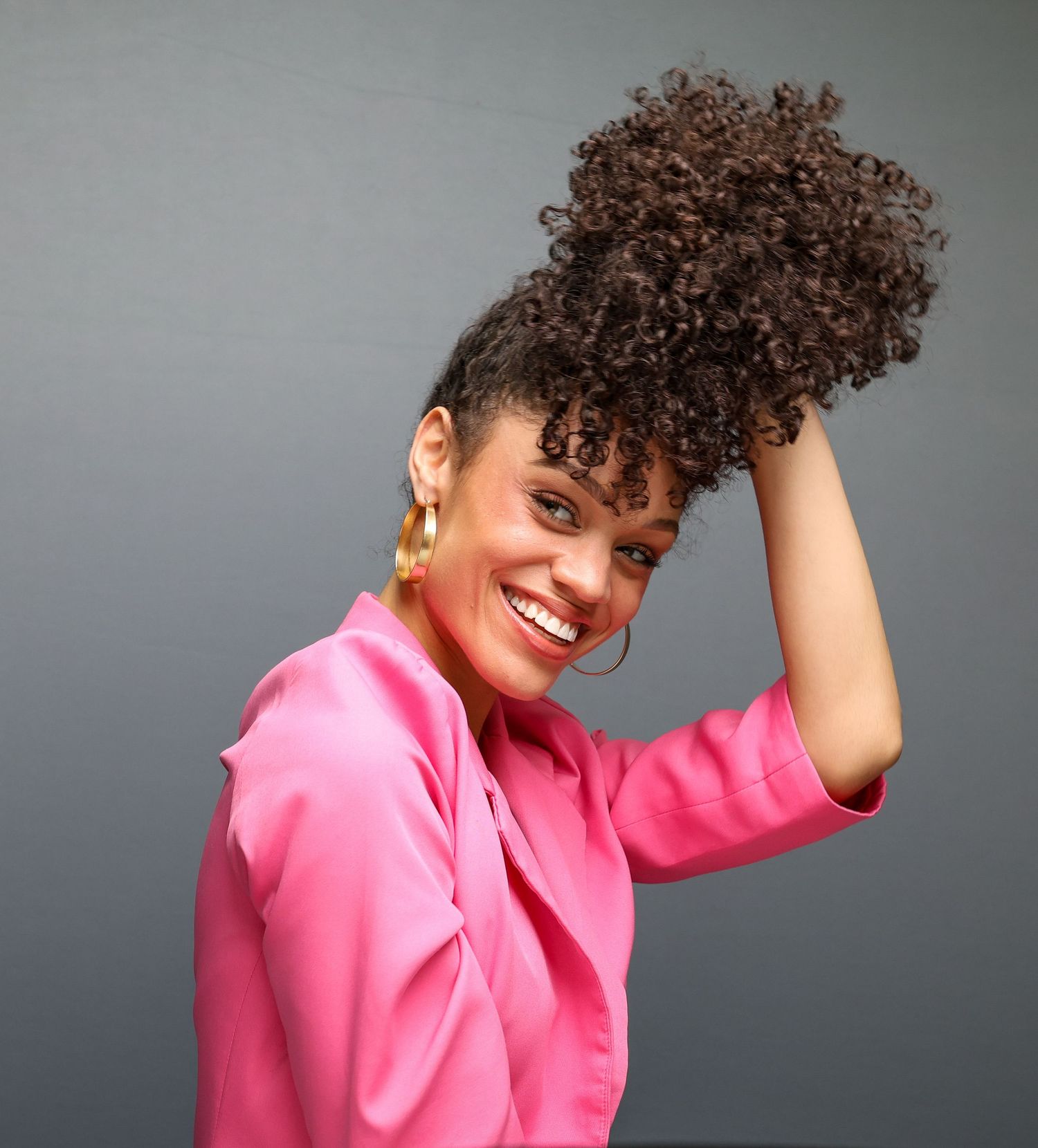 Joyful model in pink blazer with natural curly updo hairstyle poses against gray background.
