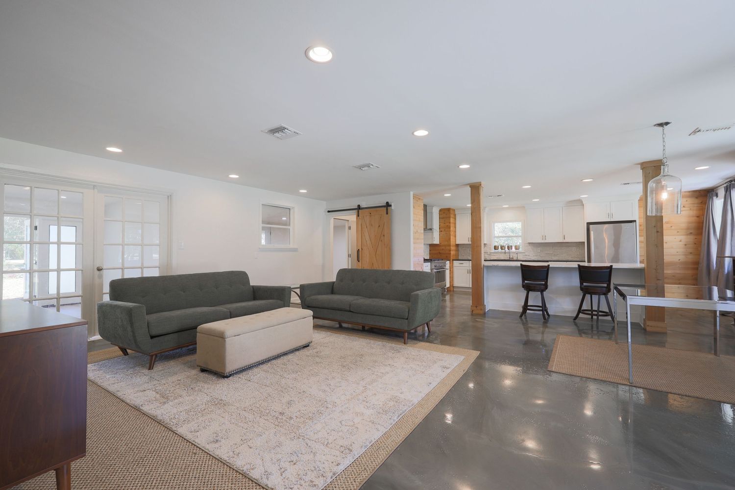 Modern open concept living room with gray sofas, white ottoman and hardwood floors connecting to kitchen with bar seating.