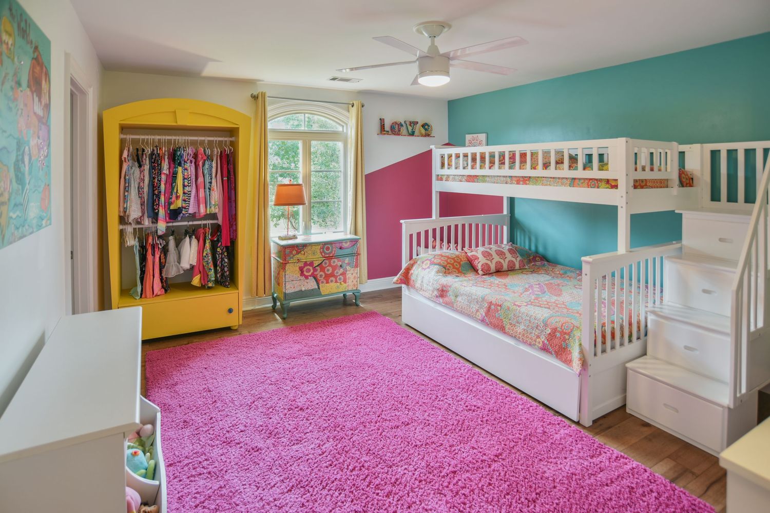 Colorful children's bedroom with white bunk bed, pink carpet, teal walls, and yellow wardrobe filled with clothes.