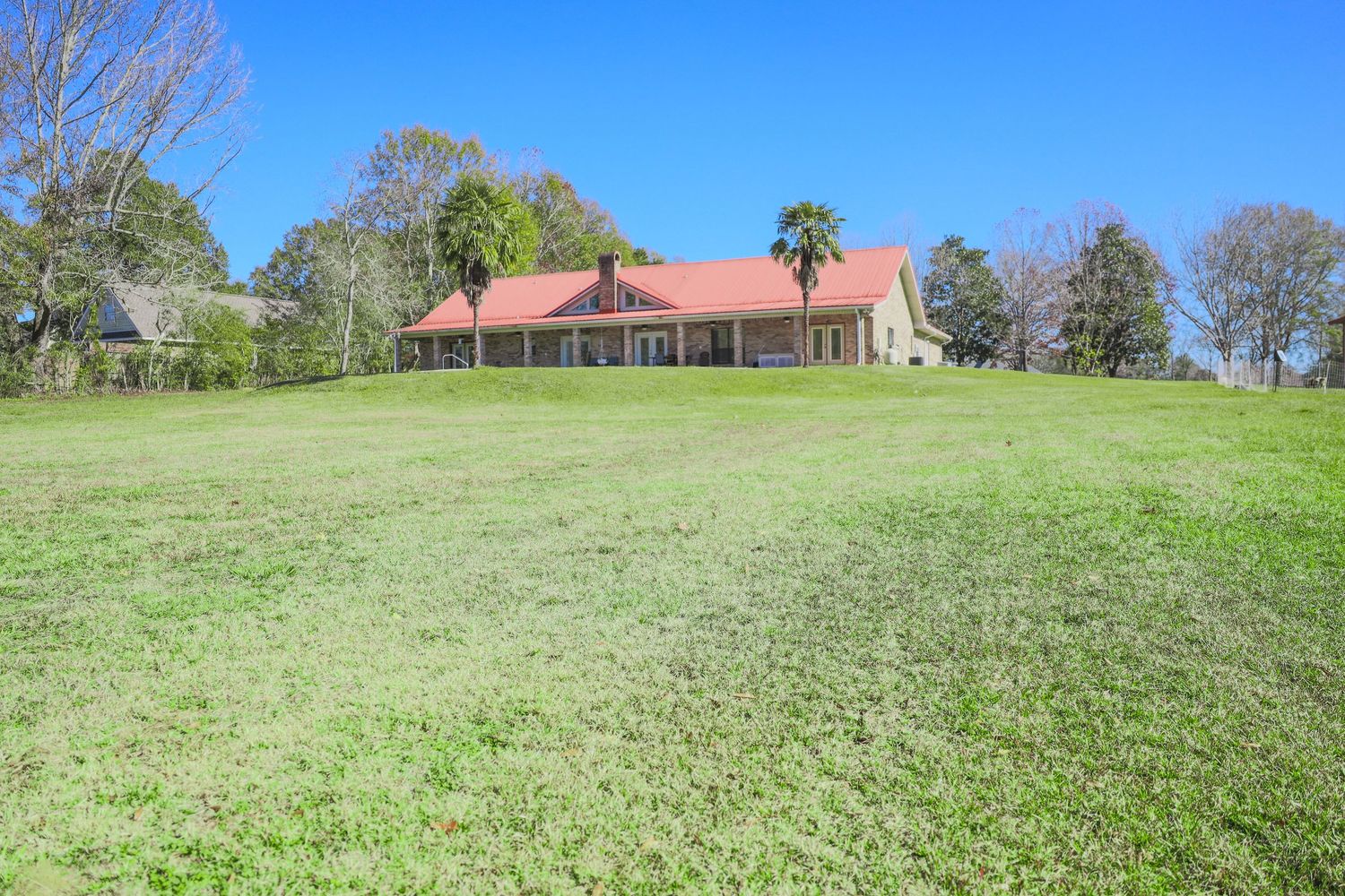 A rustic ranch house with terracotta roof sits atop a sprawling green hill surrounded by trees on a sunny day.