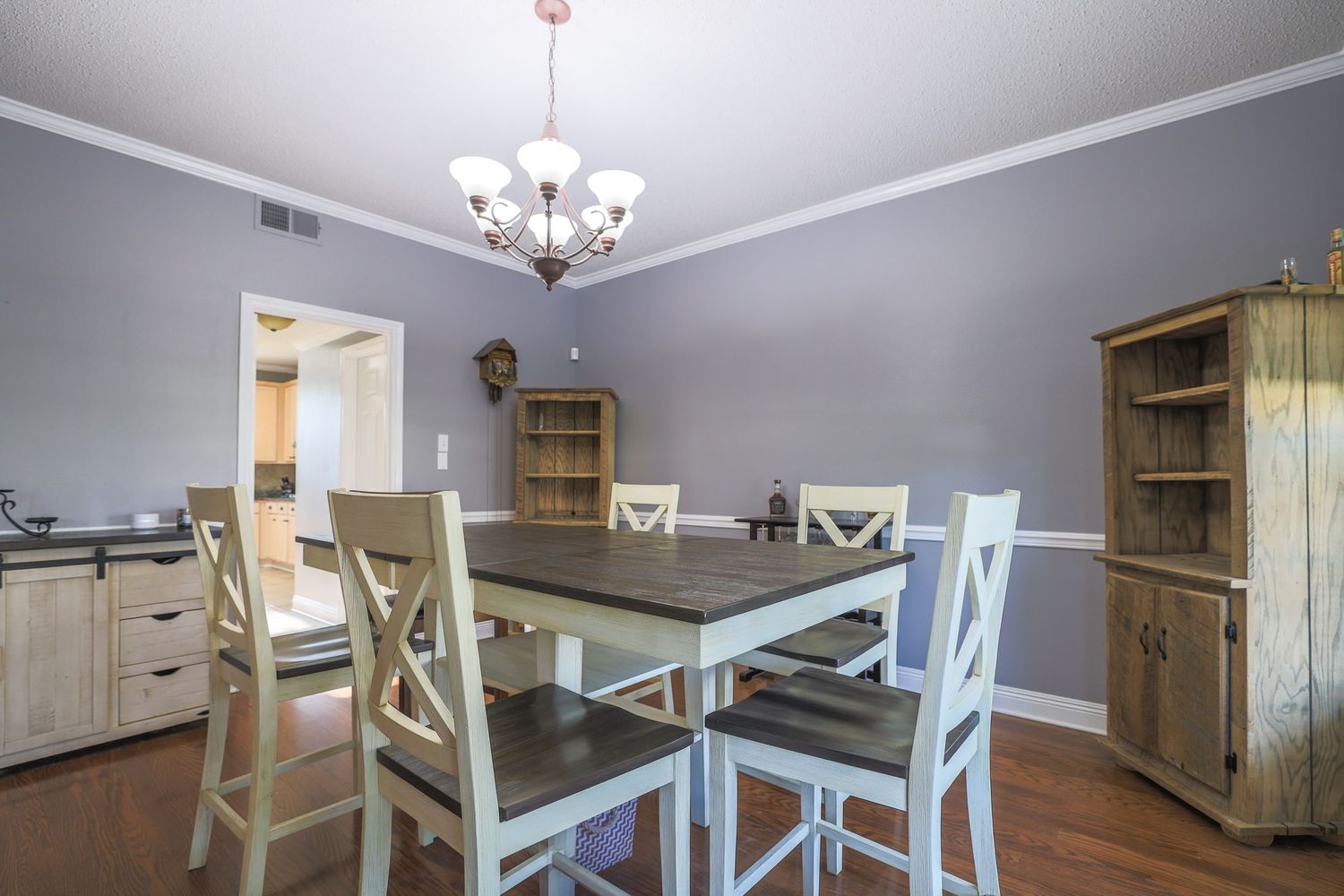 Modern dining room with gray walls, white chairs around wooden table, and rustic corner cabinets under chandelier lighting.