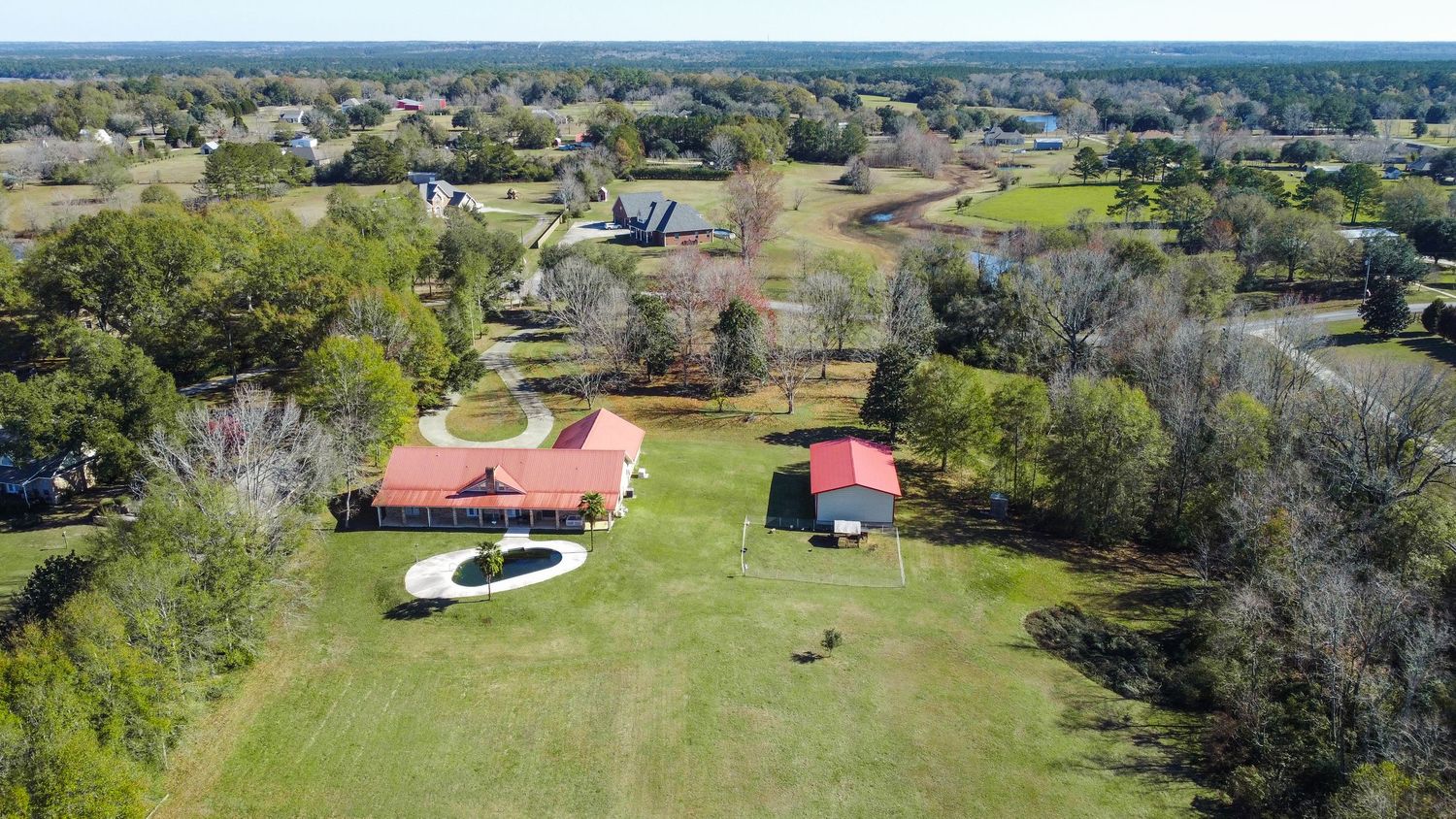 Aerial view of rural property with red-roofed buildings, swimming pool, and expansive green lawn surrounded by trees.