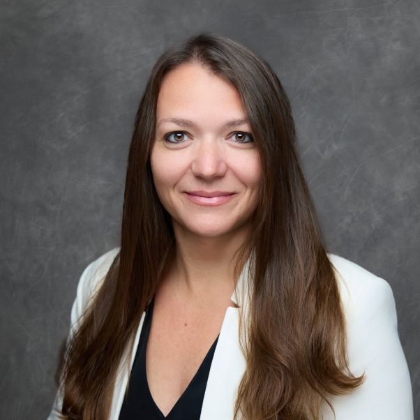 Professional portrait of person in white blazer and black top with long brown hair against gray backdrop.