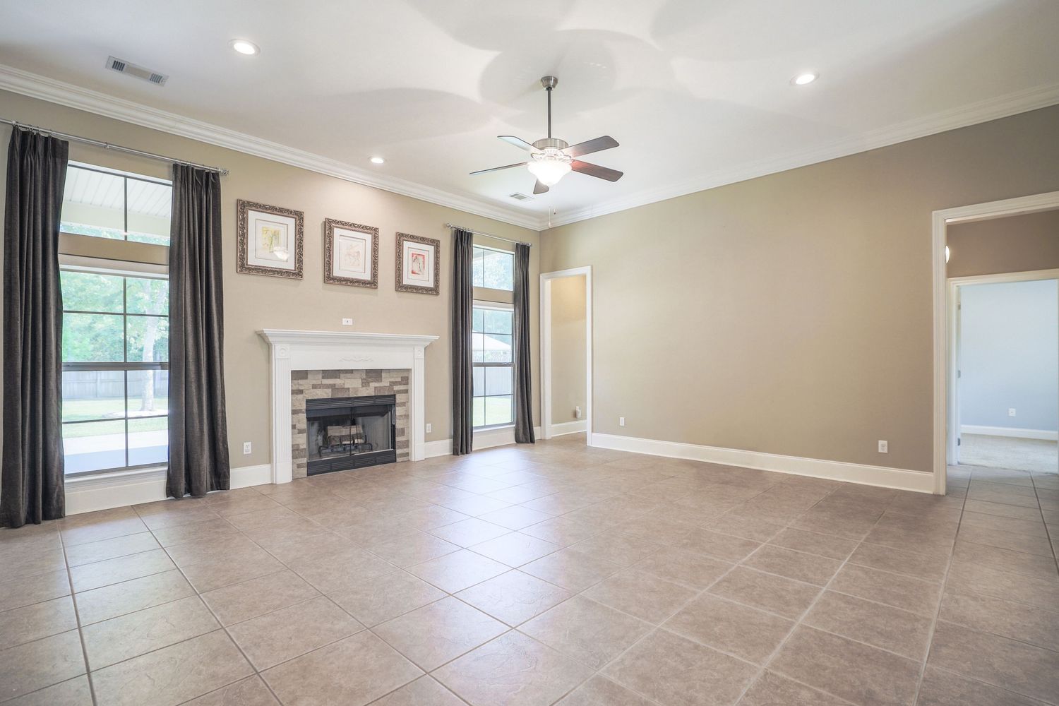 Spacious living room with fireplace, crown molding, beige walls, tile flooring and tall windows with dark curtains.