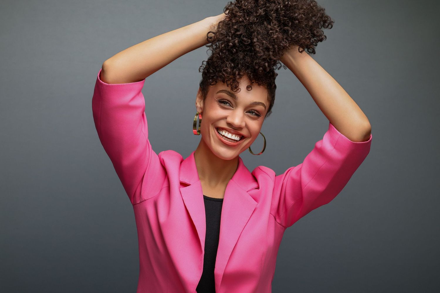 A joyful person wearing a bright pink blazer poses with hands in curly hair against a gray background.