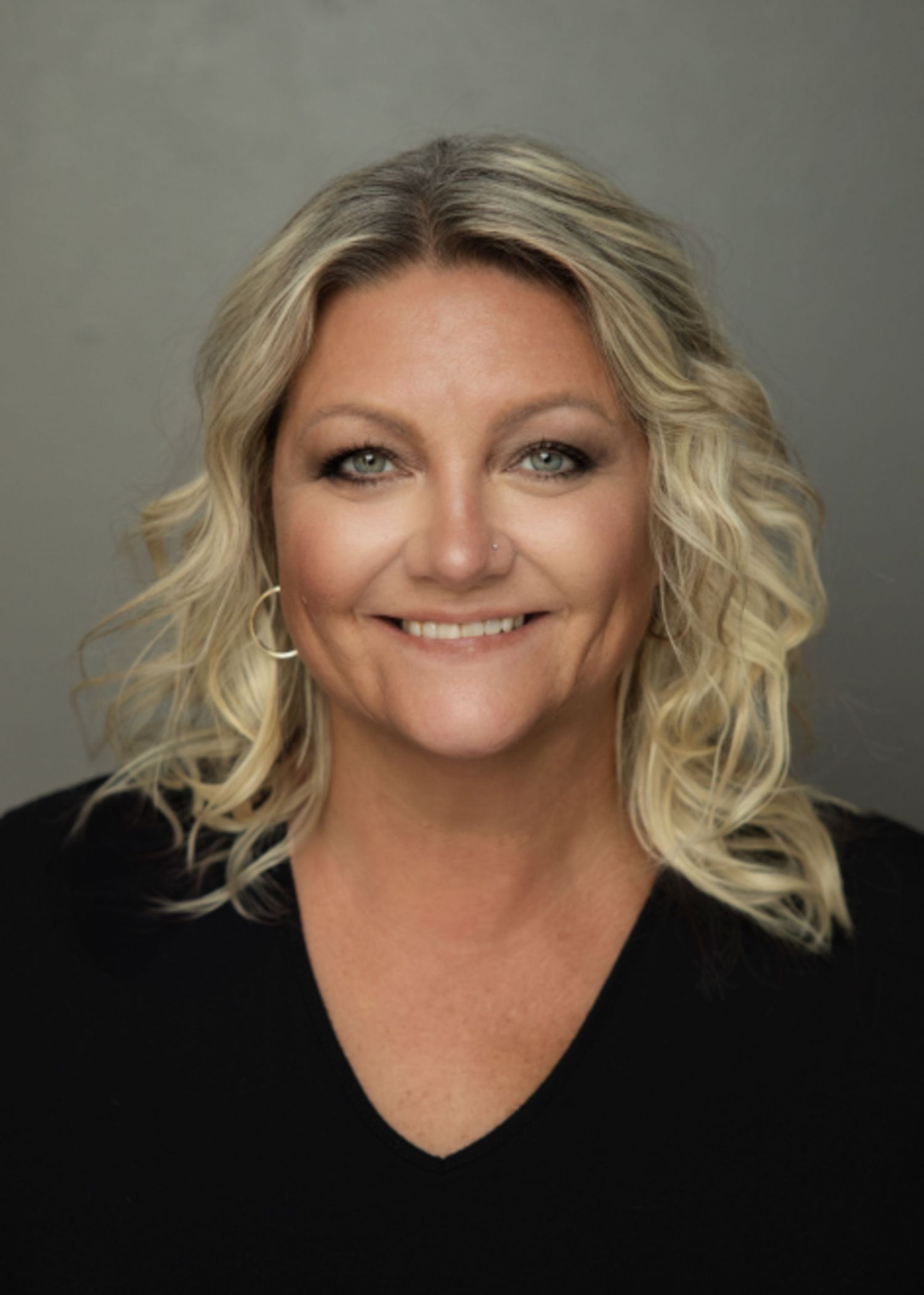 Professional headshot of someone with wavy blonde hair wearing a black V-neck top against a neutral gray background.