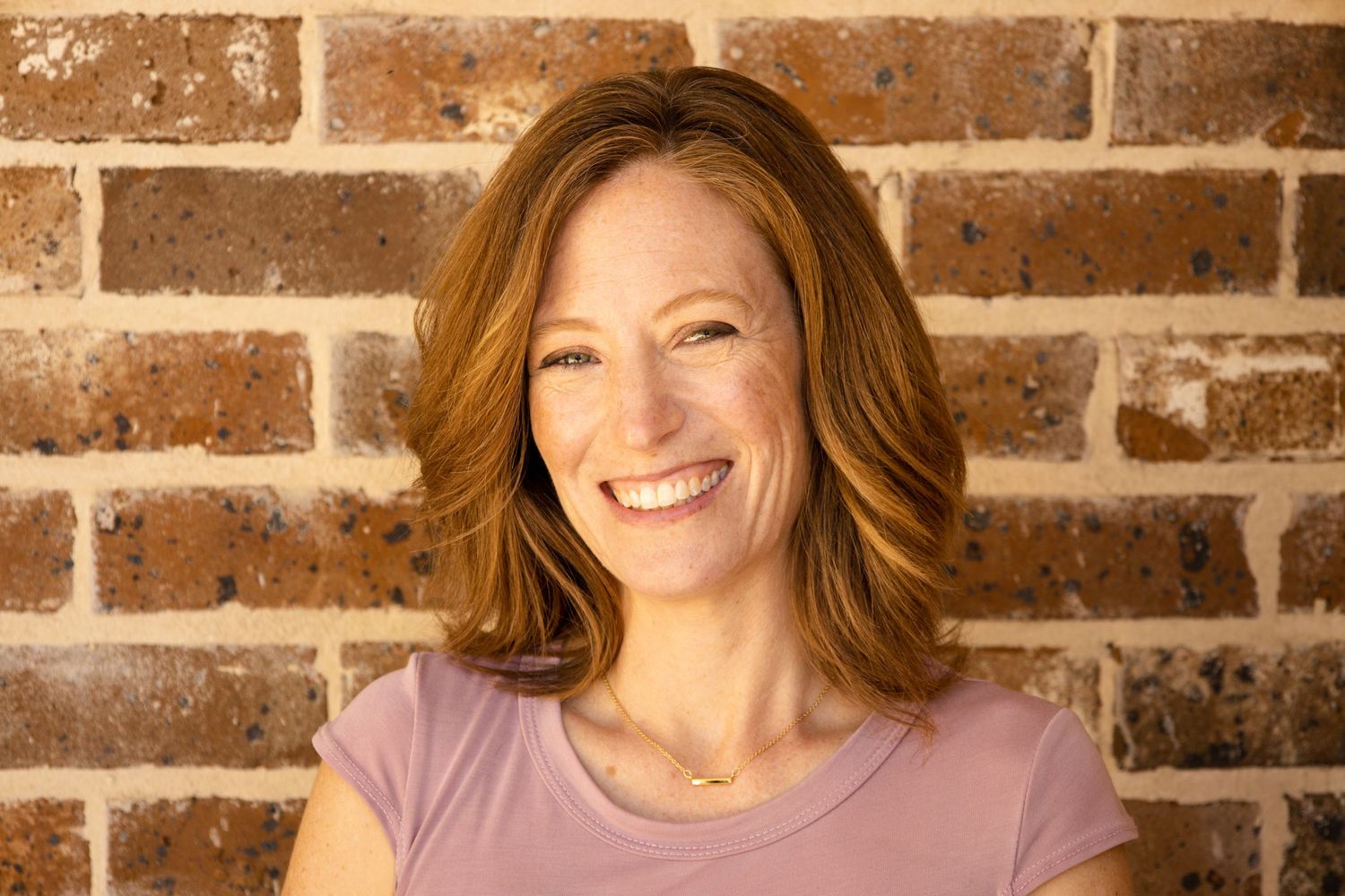 A headshot with a cheerful expression radiates warmth against a rustic brick wall backdrop, wearing a mauve colored top.