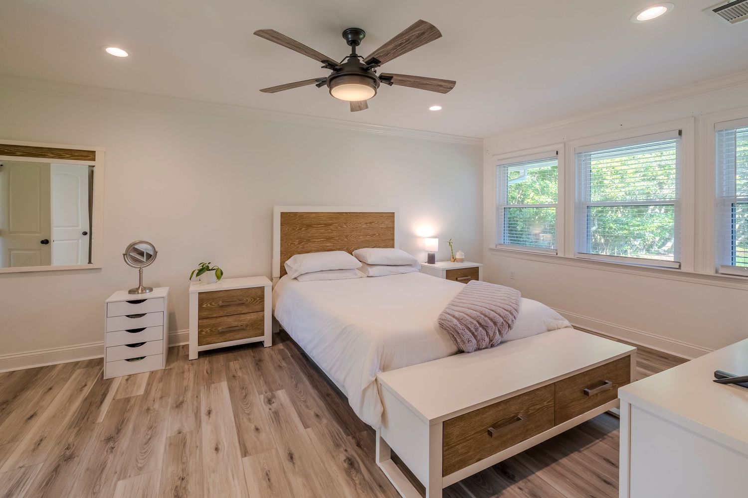 Modern bedroom with wood flooring, ceiling fan, rustic headboard and white storage furniture next to bright windows.