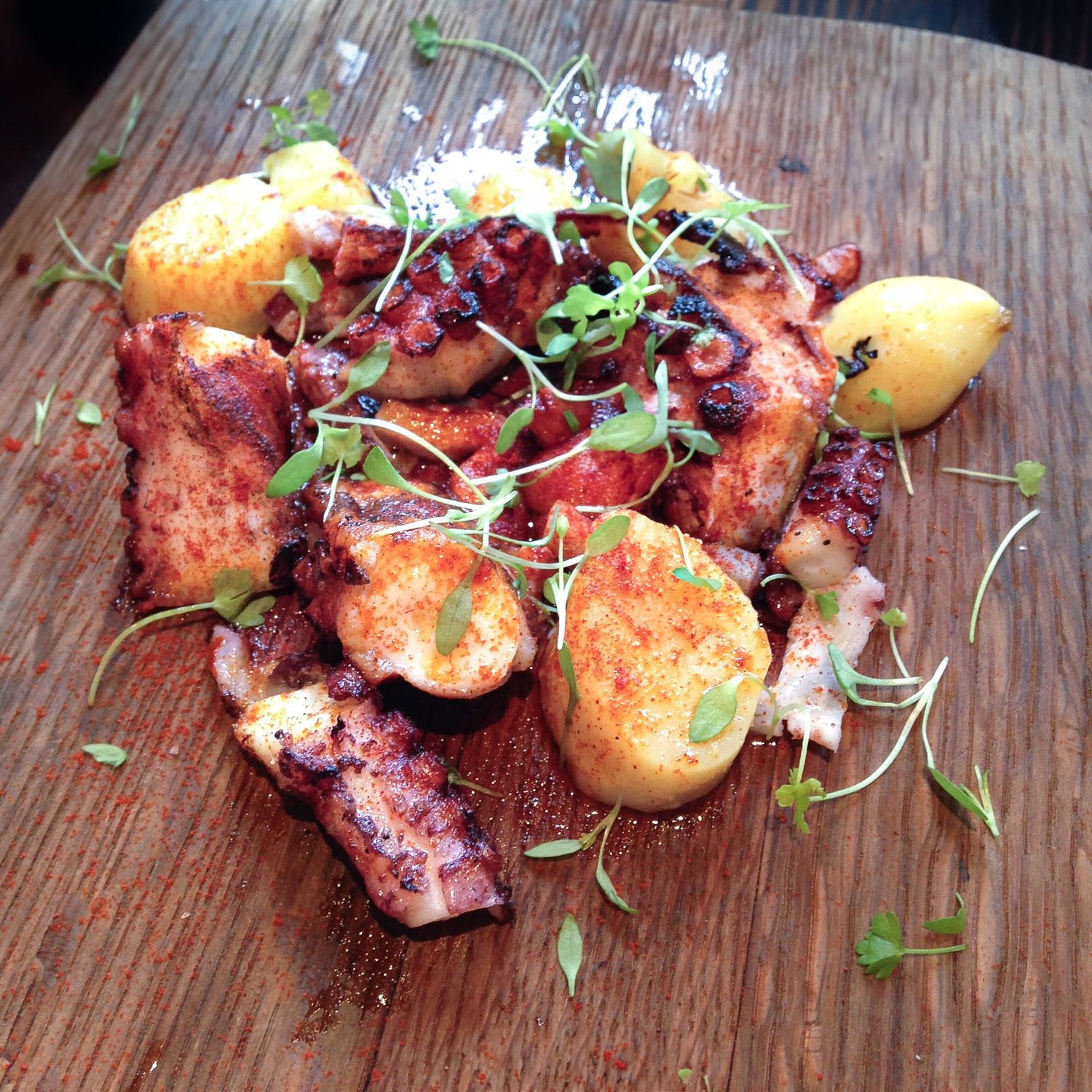 Grilled octopus dish with crispy potatoes and micro greens served on a rustic wooden board.