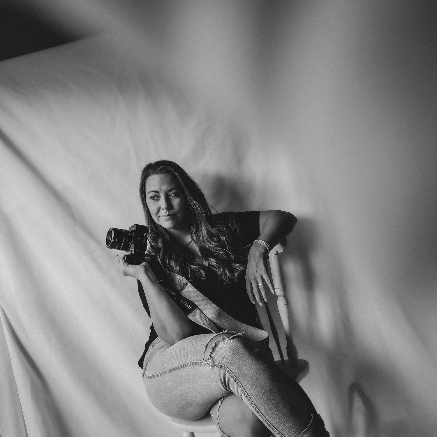 Black and white photo of a smiling woman sitting on a stool holding a camera against a draped fabric backdrop.