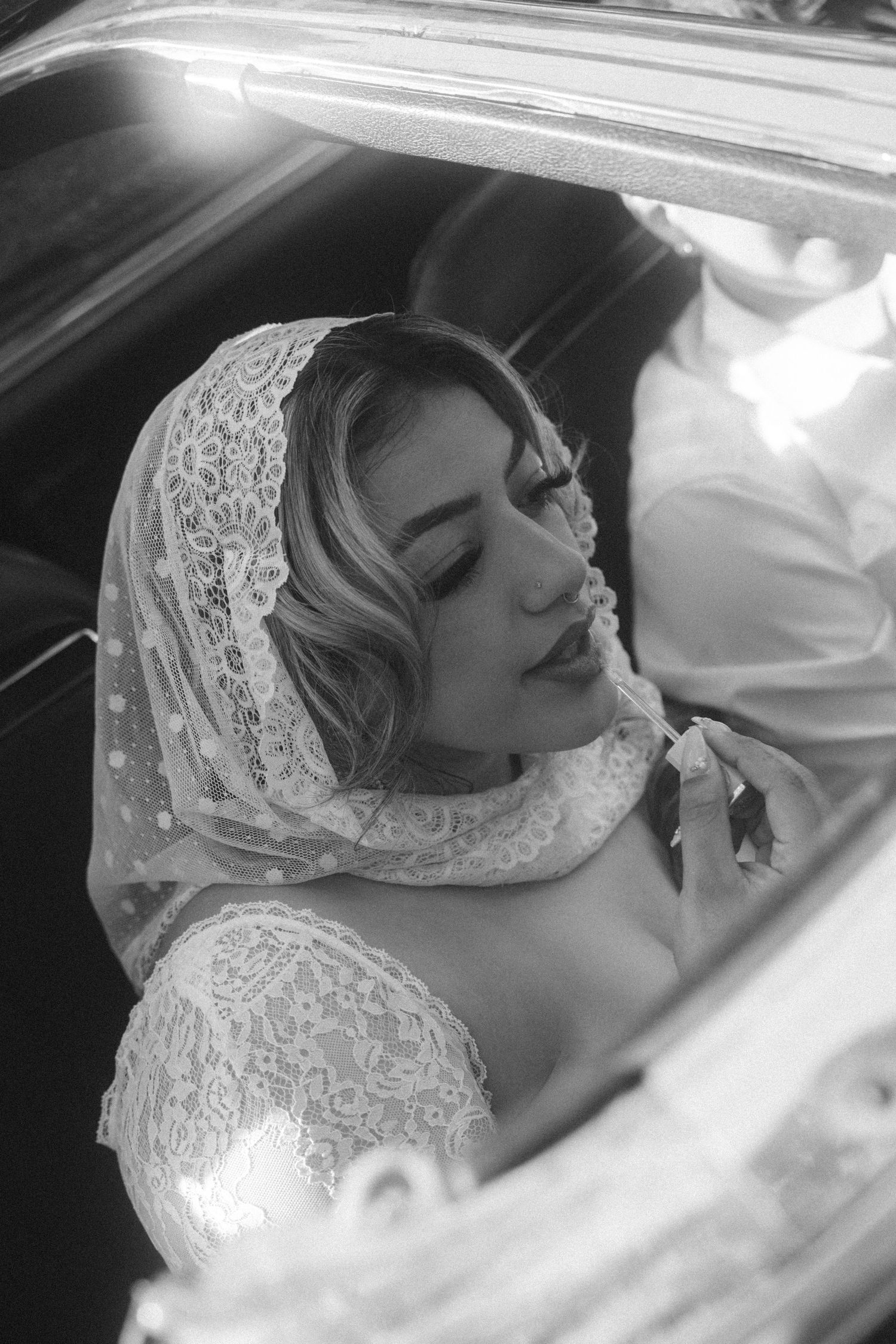Elegant black and white portrait of someone in lace wedding attire resting in a car.