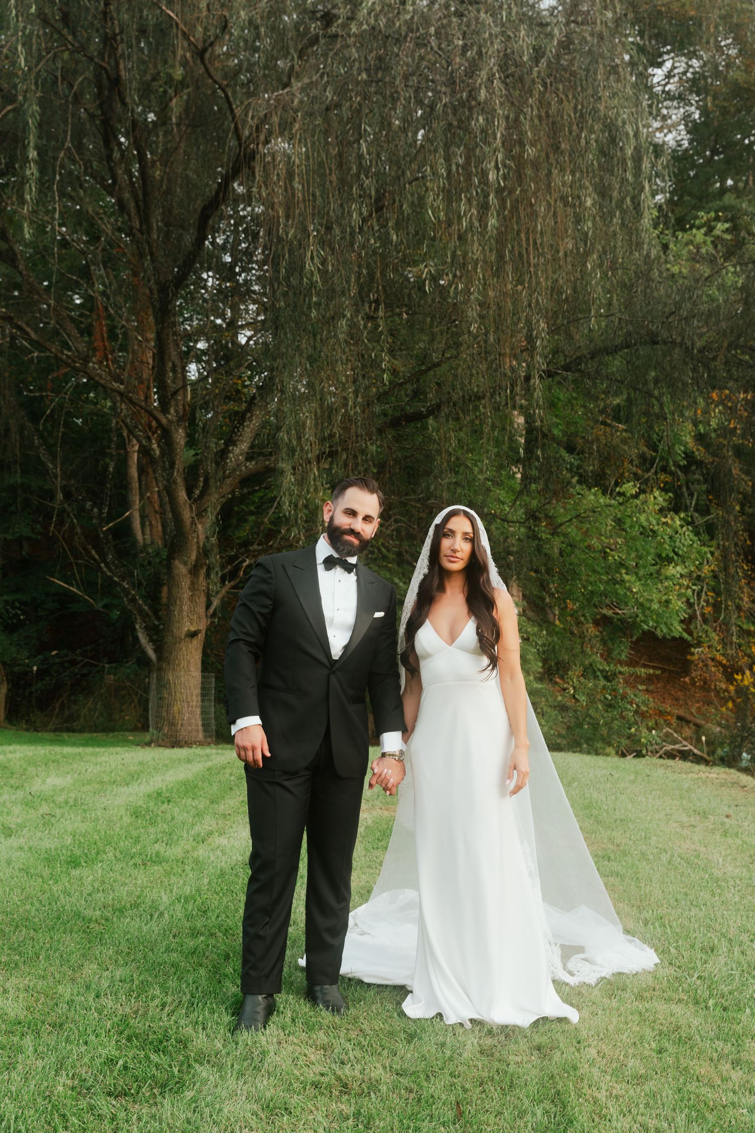Bride and groom holding hands on green lawn with dramatic weeping willow tree backdrop on wedding day.