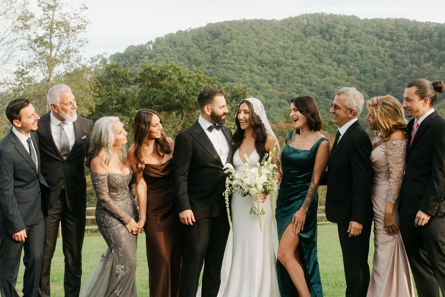 Wedding party in formal attire laughing outdoors with lush green mountains in the background.