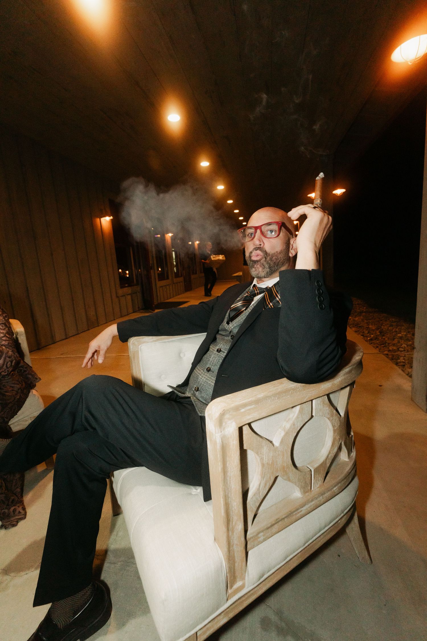 Bald man in black suit smoking a cigar while relaxing in a white armchair at an evening event.