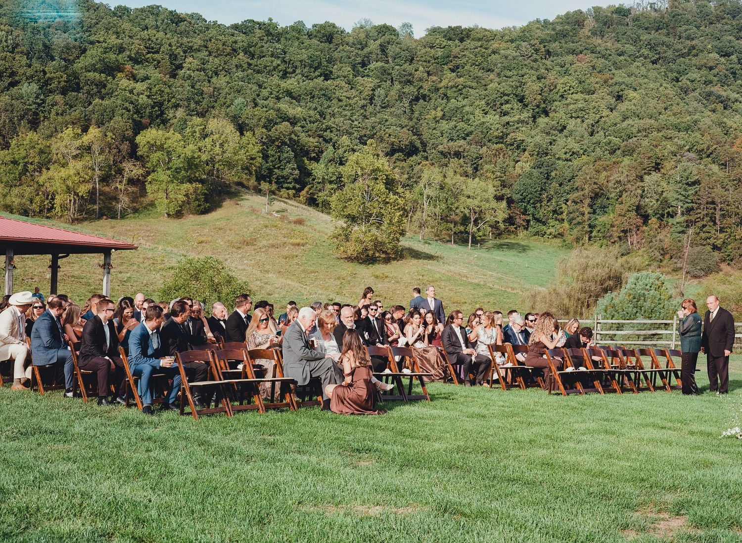 Wedding guests seated in wooden chairs on green hillside lawn for outdoor ceremony surrounded by lush forest.